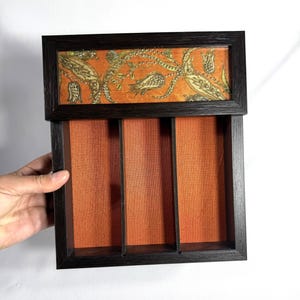 May include: A rectangular wooden organizer with a dark brown frame and three vertical compartments. The interior base is lined with a solid orange fabric. The top section features a decorative panel with a gold and orange floral pattern.
