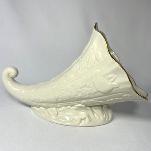 May include: Cream-colored cornucopia-shaped decorative piece. The ceramic horn of plenty features embossed fruit and foliage designs. The rim is trimmed with gold. The base is also cream-colored and has a leaf design.