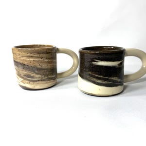 May include: Two handmade ceramic mugs. One mug features a marbled pattern in shades of brown and tan. The other mug has a dark brown and white design. Both mugs have a beige handle.
