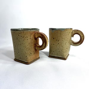 May include: Two handmade ceramic mugs with a square shape and rounded handles. The mugs have a speckled, earthy tone with a brown base and rim. The handles are a darker brown. The mugs are displayed on a white surface.