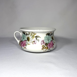 May include: A white ceramic bowl with a handle, adorned with a floral pattern featuring pink, teal, and yellow flowers. The bowl has a slightly flared rim and a small handle on one side, perfect for serving or display.