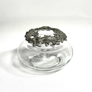 May include: A clear glass bowl with a decorative metal floral rim. The rim features detailed, three-dimensional flowers and leaves. The bowl has a rounded shape and sits on a white surface. The metal rim is a dark silver color.