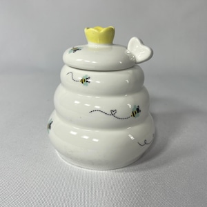 May include: White ceramic honey pot shaped like a beehive, with a yellow crown on top and a butterfly-shaped handle. The pot is decorated with small bees and dotted lines. A charming addition to any kitchen.