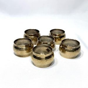 May include: Six small, gold-colored metal bowls with a hammered texture. The bowls are arranged in a circular pattern, each with a rounded shape and a slightly flared rim. The bowls are likely for decorative or serving purposes.