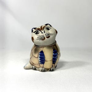May include: A ceramic cat figurine with a cream-colored head and a body featuring gold and blue accents. The cat's face has brown markings and black details. The figurine has a silver-colored collar.