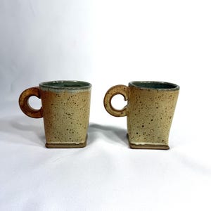 May include: Two handmade ceramic mugs with a square base and a rounded handle. The mugs have a speckled tan exterior, a dark green interior, and a unique, organic shape. The mugs are perfect for coffee or tea.
