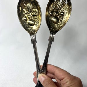 May include: A pair of vintage serving spoons with ornate gold-toned bowls and silver-toned handles. The bowls feature a detailed embossed design of fruits and foliage. The handles have a decorative, scalloped design at the base.