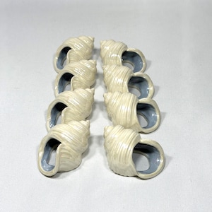 May include: Eight ceramic seashell napkin rings. The rings are off-white with a spiral design and a light blue interior. The napkin rings are arranged in two columns of four.