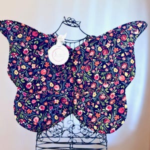 May include: Butterfly-shaped fabric wings with a dark blue background and a floral pattern in shades of pink, yellow, and orange. The wings are displayed on a black wire mannequin. A white tag with a logo is attached.