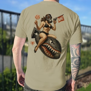 May include: Tan t-shirt featuring a pin-up girl wearing a gas mask and bikini, riding a bomb with a shark mouth design. The girl holds a lollipop and a gun with a "BANG!" sign. The shirt has a vintage, military-inspired aesthetic.