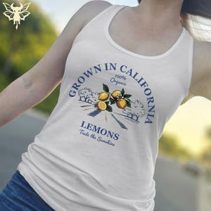 Puede incluir: Camiseta sin mangas blanca con un gráfico que dice "Grown in California" alrededor de una ilustración de limón. El texto "100% Organic" está encima de la ilustración, y "Lemons Taste the Sunshine" debajo.