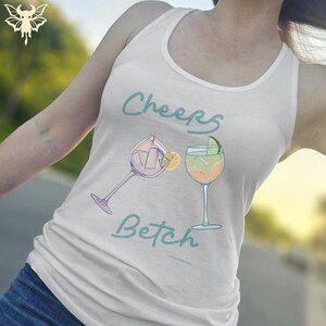 May include: White tank top with the words "Cheers Betch" in teal script, along with illustrations of two cocktails. One cocktail is purple with a lemon slice, the other is green and orange with a lime wedge.