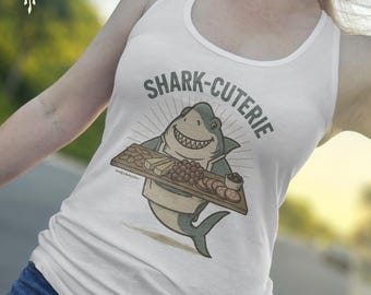 May include: White tank top featuring a cartoon shark holding a charcuterie board. The shark is gray and white, wearing a chef's apron, and has a big smile. The text "SHARK-CUTERIE" is above the shark in gray letters.