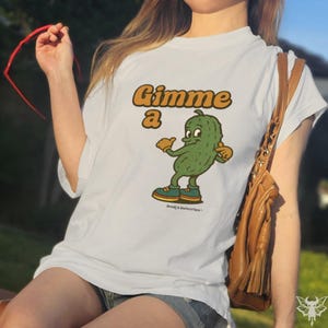 May include: White t-shirt with a cartoon pickle graphic. The pickle is green with orange gloves and shoes, and the text "Gimme a" in orange. The shirt is a casual style.