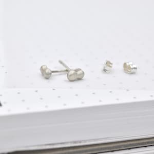 May include: A pair of silver stud earrings, each featuring two small, rounded spheres joined together in a linear design. The earrings include matching silver butterfly-style earring backs, all displayed with a matte, textured metallic finish.