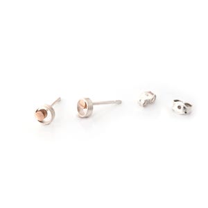 May include: Silver stud earrings with a rose gold disc detail. The earrings are shown with two butterfly backs. The design is minimalist and modern. The earrings are suitable for everyday wear.