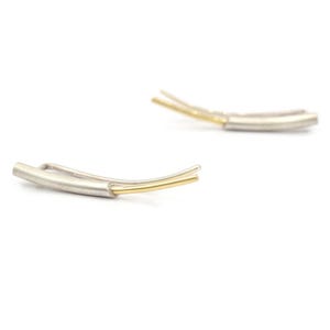 May include: A pair of silver and gold earrings with a curved design. The earrings are made of two thin metal bars, one silver and one gold, that are joined together at one end. The earrings are designed to be worn on the earlobe.