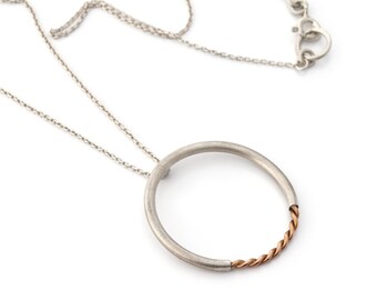 May include: Silver-tone necklace featuring a large, open circle pendant. The pendant has a brushed metal finish with a section of twisted copper-colored metal. The necklace has a delicate chain and a secure clasp.