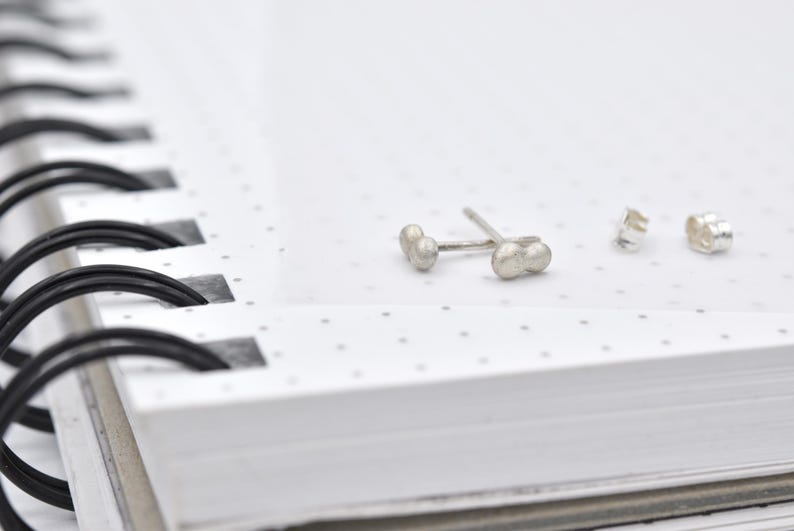 K&ouml;nnte beinhalten: Ein Paar Ohrstecker aus Silber, bei denen jeweils zwei kleine, abgerundete Kugeln in einem linearen Design miteinander verbunden sind. Die Ohrstecker werden mit passenden silbernen Schmetterlingsverschl&uuml;ssen geliefert und haben eine matte, texturierte Oberfl&auml;che.