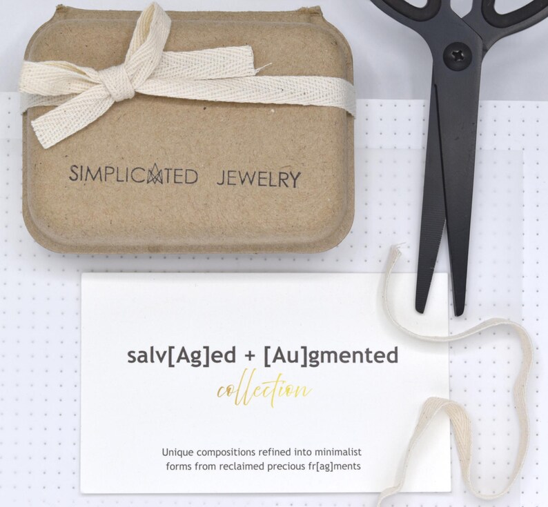 K&ouml;nnte beinhalten: Eine beigefarbene Schmuckschachtel aus Karton mit einer cremefarbenen Schleife, einer schwarzen Schere und einer wei&szlig;en Karte. Die Schachtel ist mit SIMPLICATED JEWELRY bedruckt. Die Karte zeigt den Text salv[Ag]ed + [Au]gmented collection.