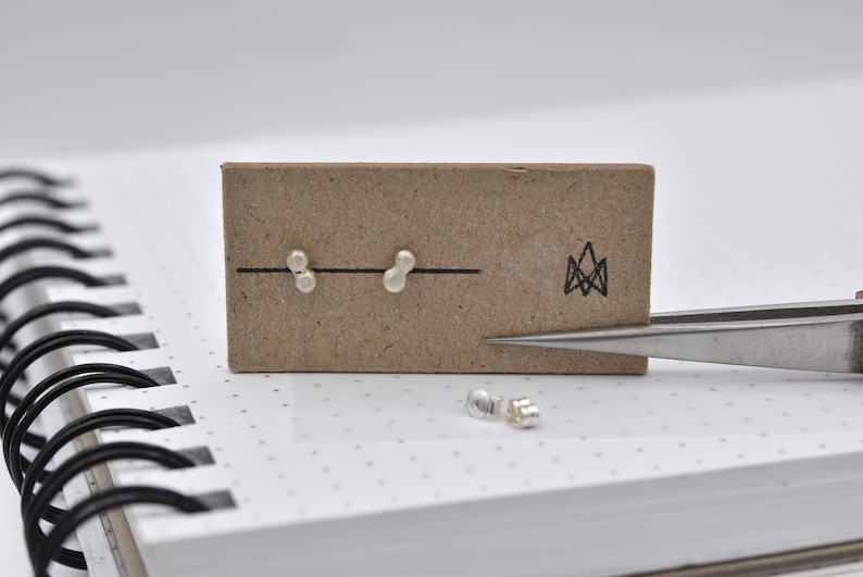 K&ouml;nnte beinhalten: Ein Paar kleine silberne Ohrstecker, die jeweils aus zwei verbundenen Kugeln bestehen, befestigt auf einer rechteckigen braunen Pappkarte. Die Karte zeigt ein schwarzes geometrisches Logo und eine horizontale Linie. Zwei silberne Ohrstecker-Verschl&uuml;sse liegen daneben.