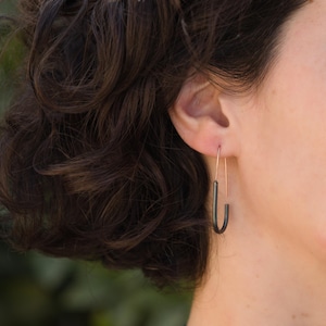 May include: A close-up shot of a person's ear wearing a pair of modern, minimalist earrings. The earrings feature a slender, dark gray, U-shaped pendant suspended from a thin, copper-colored wire. The design is simple and elegant.