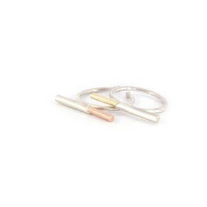 May include: A collection of silver rings with bar and band designs. One ring features a gold bar, another a rose gold bar, and the others are simple silver bands. The jewelry is set against a white background.