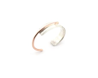 Könnte beinhalten: Ein silberner Ring mit einem roségoldenen Akzent. Der offene Ring hat einen gebogenen roségoldenen Stab, der sich über die Oberseite erstreckt. Der Schmuck ist vor einem weißen Hintergrund platziert.
