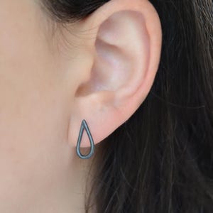 Peut inclure: Gros plan sur une oreille portant une boucle d'oreille unique en forme de goutte d'eau gris foncé. Le bijou a un design minimaliste. L'arrière-plan est flou, mettant en valeur la forme de la boucle d'oreille.