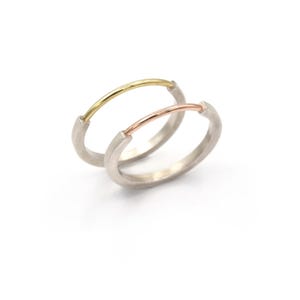 May include: Two minimalist rings. One ring features a gold-colored bar across a silver band. The other ring has a rose gold-colored bar across a silver band. The rings are set against a white background.