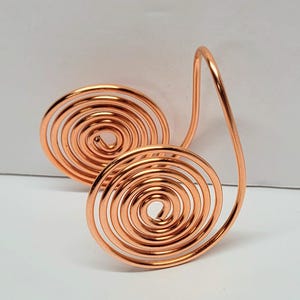 May include: A copper-colored metal wire sculpture featuring two spiral shapes connected by a curved line. The spirals are tightly wound, and the overall design is symmetrical. The sculpture is displayed against a white background.