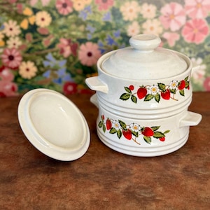 Puede incluir: Una pila de dos ollas de cerámica color hueso con tapas, texturas moteadas y asas laterales. Cada olla presenta un patrón decorativo de fresas rojas, hojas verdes y flores blancas. Una tapa a juego se encuentra colocada por separado a la izquierda.