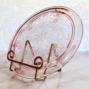 May include: Pink glass oval serving platter with embossed floral design, displayed on a gold-colored metal stand. The platter features a detailed pattern of daisies and leaves. A decorative piece for serving or display.