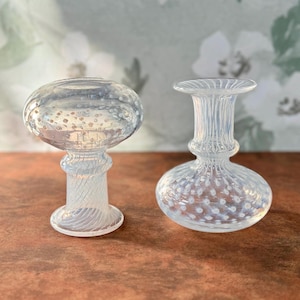 Op de afbeelding: Twee decoratieve objecten van helder glas. Het object links heeft een bolvormige bovenkant met kleine belletjes en een getextureerde steel. Het object rechts heeft een ronde basis, een getextureerde steel en een uitlopende bovenkant. Beide objecten staan op een bruin oppervlak.