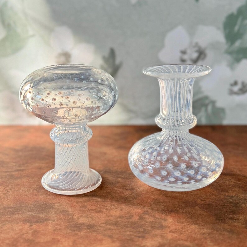 Op de afbeelding: Twee decoratieve objecten van helder glas. Het object links heeft een bolvormige bovenkant met kleine belletjes en een getextureerde steel. Het object rechts heeft een ronde basis, een getextureerde steel en een uitlopende bovenkant. Beide objecten staan op een bruin oppervlak.