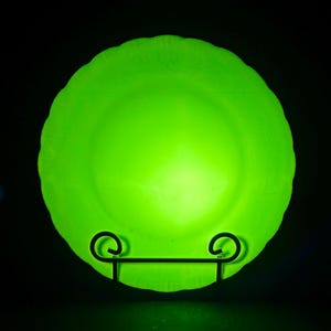 May include: A bright green glass plate with a scalloped edge, displayed on a black metal stand. The plate has a textured surface and is illuminated against a dark background. The plate is round and appears to be a decorative or serving piece.