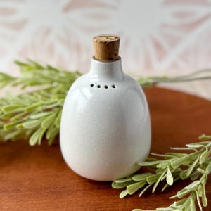 May include: A small, rounded, off-white ceramic shaker with a smooth, speckled glaze finish. The top features a narrow neck fitted with a light brown cylindrical cork stopper. Four small, evenly spaced holes are visible on the upper shoulder.