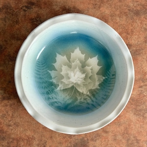 May include: A round ceramic pie dish with a scalloped edge, featuring a white speckled glaze on the rim and a gradient blue interior. The center of the dish displays a large, detailed white maple leaf imprint, flanked by delicate fern leaf patterns.