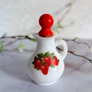 May include: A small white milk glass pitcher featuring a colorful strawberry graphic on the front. The pitcher has a curved handle and a spout, and it is topped with a bright red, spherical stopper that fits securely into the opening.