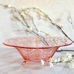 Könnte beinhalten: Eine rosa Glasschale mit einem dekorativen floralen Muster. Die Schale hat ein breites, flaches Design und wird von drei kleinen Füßen getragen. Die Kanten sind leicht gewellt. Die Schale ist wahrscheinlich Vintage oder antik.