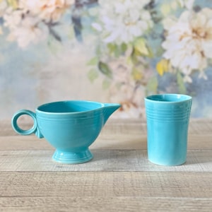 Può includere: Una lattiera in ceramica turchese con manico e beccuccio, e un bicchiere abbinato. La lattiera ha un manico rotondo e poggia su una base a piedistallo. Il bicchiere presenta delle scanalature orizzontali. Entrambi sono su una superficie di legno.