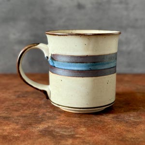 May include: Cream-colored ceramic mug with a brown handle and rim. The mug features two horizontal bands: a dark brown band above a light blue band. The mug is a simple, yet elegant design.