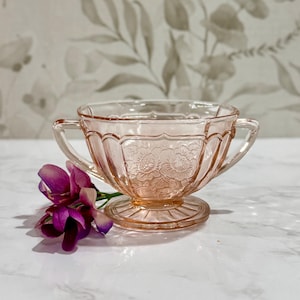 May include: A vintage pink glass sugar bowl with two handles and a floral design. The bowl is on a white surface with a purple flower.