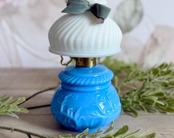 Puede incluir: Una lámpara de aceite en miniatura con una base de vidrio azul brillante con patrones florales en relieve y una pantalla de vidrio lechoso blanco con nervaduras verticales. Un pequeño lazo de tela gris está atado a la parte superior de la pantalla, con un cuello de metal.