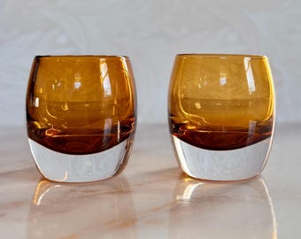 Könnte beinhalten: Zwei abgerundete Glasbecher mit einem tief bernsteinfarbenen oberen Teil, der in einen klaren, dicken Glasboden übergeht. Die Gefäße haben eine glatte, geschwungene Silhouette mit einer weiten Öffnung und einer schweren, massiven Bodenkonstruktion.