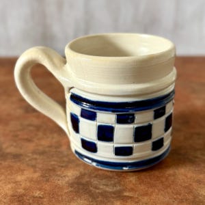 Puede incluir: Una taza de cerámica blanca con un asa negra y el texto "But First, Coffee" en negrita negra. La taza se encuentra sobre un fondo neutro, resaltando su diseño sencillo.