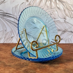 Pode incluir: Dois pratos de vidro azul claro com borda franzida são exibidos em um suporte de metal dourado. Os pratos são empilhados, com um repousando no suporte e o outro inclinado acima. O fundo apresenta um padrão floral.