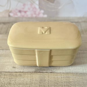 Może przedstawiać: Prostokątny, jasnożółty ceramiczny pojemnik z pokrywką. Pokrywka ma podniesiony wzór "M". Pojemnik ma poziome żłobienia i pionowy element przypominający uchwyt z przodu. Prawdopodobnie mydelniczka lub pudełko na drobiazgi.