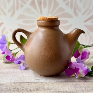 Puede incluir: Una tetera redonda de cerámica marrón con un acabado liso y mate, un asa curva, un pico corto y un tapón de corcho cilíndrico. La tetera está rodeada de flores de guisante de olor moradas y blancas con tallos y hojas verdes.