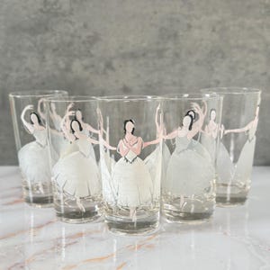 May include: A set of five clear drinking glasses, each decorated with a ballerina design. The glasses feature white ballerina illustrations with pink accents. The glasses are cylindrical and suitable for various drinks.